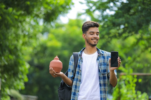 Joven universitario indio sosteniendo la hucha de arcilla en la mano sobre fondo de naturaleza