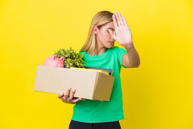 Joven uruguaya haciendo un movimiento mientras recoge una caja llena de cosas aisladas en un fondo amarillo haciendo un gesto de parada y decepcionada