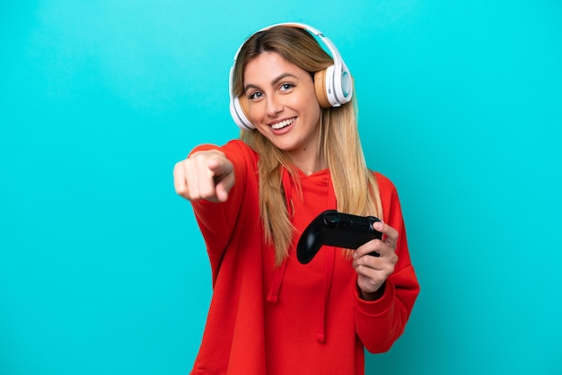 Joven uruguaya jugando con un controlador de videojuegos aislado en azul apuntando al frente con expresión feliz