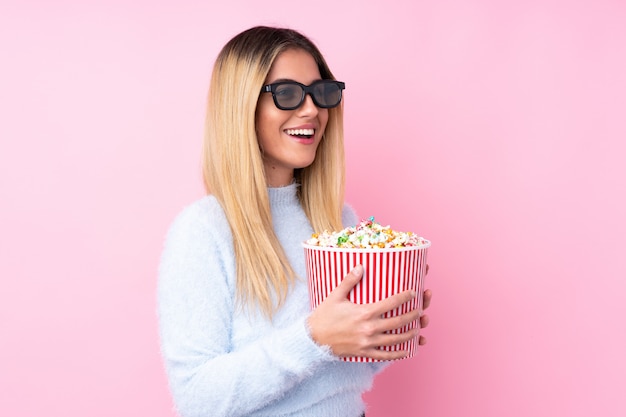 Joven uruguaya sobre pared aislada pared rosa con gafas 3d y sosteniendo un gran cubo de palomitas de maíz