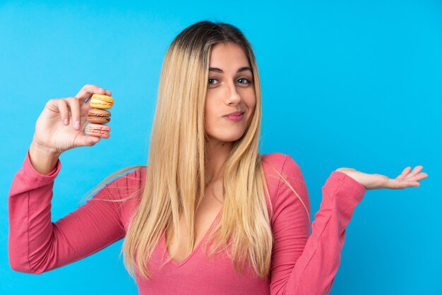 Joven uruguaya sobre pared azul aislada sosteniendo coloridos macarons franceses y haciendo gesto de dudas