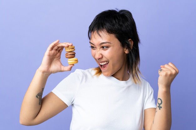 Joven uruguaya sobre pared púrpura con coloridos macarons franceses y celebrando una victoria