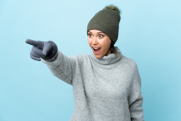 Joven uruguaya con sombrero de invierno en la pared azul apuntando lejos