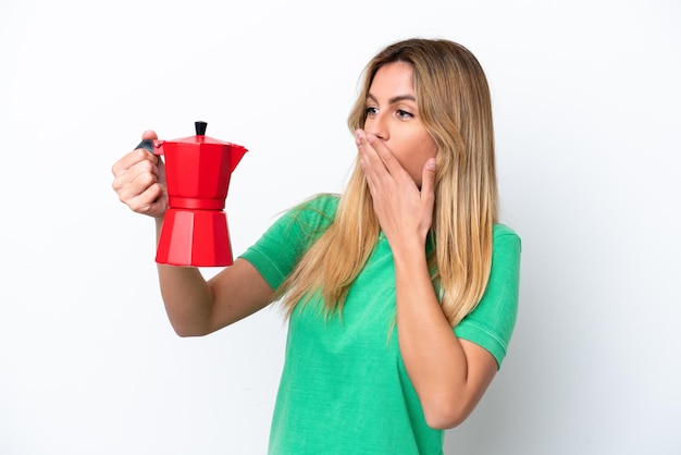 Joven uruguaya sosteniendo una cafetera aislada de fondo blanco con sorpresa y expresión facial impactada