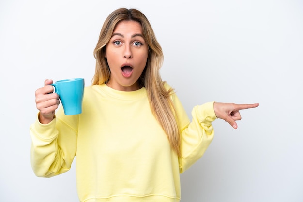 Joven uruguaya sosteniendo una taza de café aislada de fondo blanco sorprendida y apuntando hacia el lado