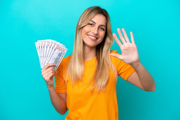 Joven uruguaya tomando mucho dinero aislada de fondo azul contando cinco con los dedos