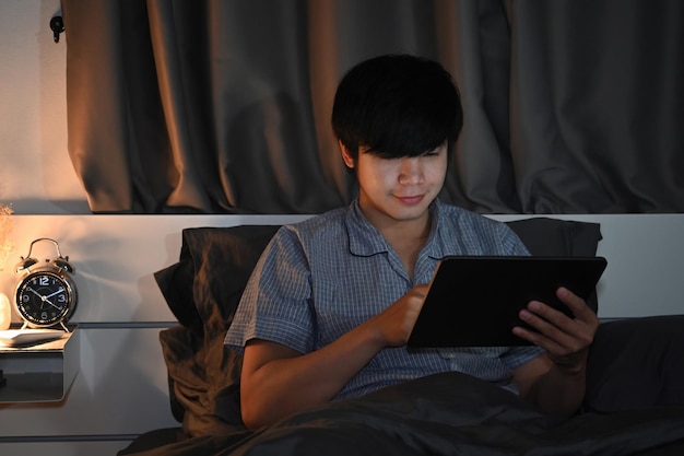 Joven usando una tableta digital mientras se sienta en la cama por la noche.