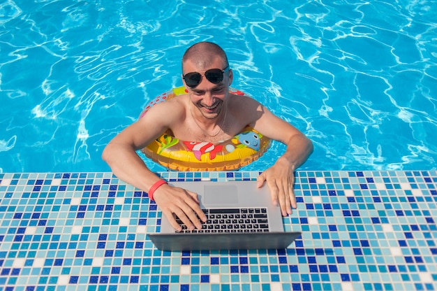 El joven en vacaciones de verano con el portátil, negocios en línea. Distancia oficina en casa en piscina en anillo de goma en cuarentena
