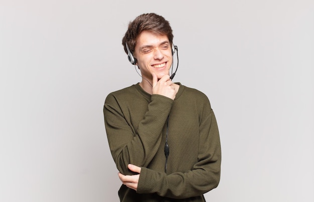 joven vendedor telefónico sonriendo con una expresión feliz y segura con la mano en la barbilla, preguntándose y mirando hacia un lado