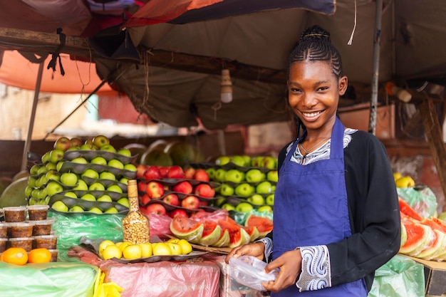 Joven vendedora de frutas africana con delantal azul de pie frente al puesto de frutas