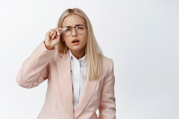 Una joven vendedora con mala visión no puede ver con gafas entrecerrando los ojos de pie sobre fondo blanco en traje