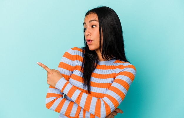 Joven venezolana aislada en la pared azul apunta con el dedo pulgar lejos, riendo y despreocupada.