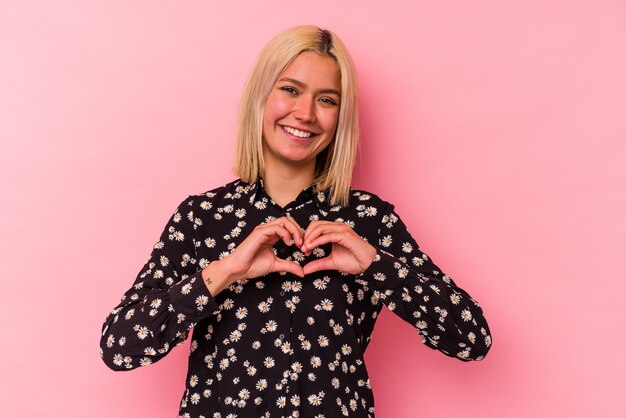 Joven venezolana aislada en la pared rosa sonriendo y mostrando una forma de corazón con las manos