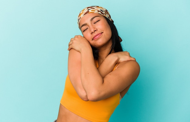 Joven venezolana aislada sobre fondo azul abrazos, sonriendo despreocupada y feliz.