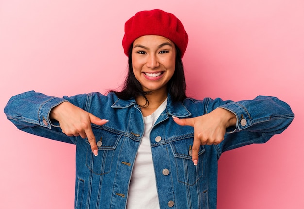 Joven venezolana aislada sobre fondo rosa apunta hacia abajo con los dedos, sentimiento positivo.