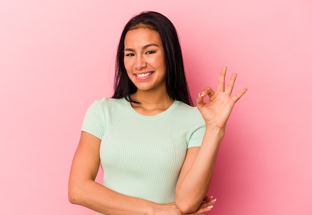 Joven venezolana aislada sobre fondo rosa guiña un ojo y tiene un buen gesto con la mano.