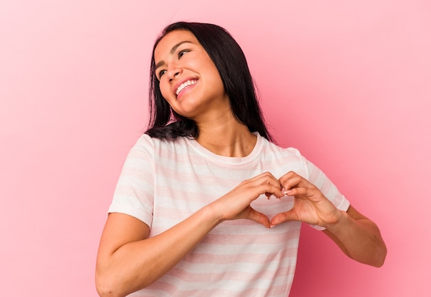 Joven venezolana aislada sobre fondo rosa sonriendo y mostrando una forma de corazón con las manos.