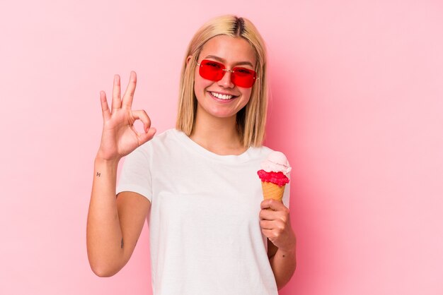 Joven venezolana comiendo un helado aislado en la pared rosa alegre y confiado mostrando gesto ok.