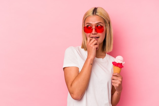 Joven venezolana comiendo un helado aislado en la pared rosa relajado pensando en algo mirando un espacio de copia.