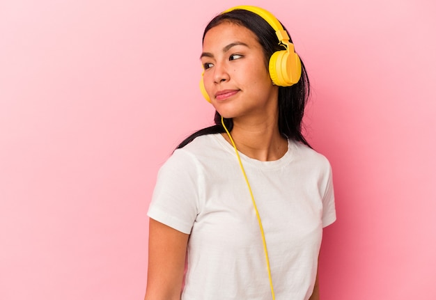 Joven venezolana escuchando música aislada sobre fondo rosa se ve a un lado sonriente, alegre y agradable.