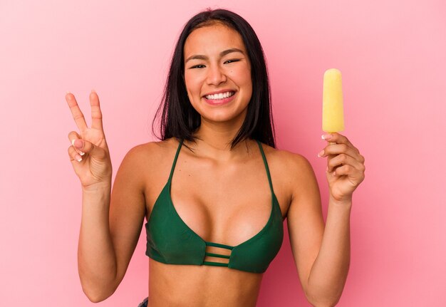 Joven venezolana sosteniendo un helado aislado sobre fondo rosa alegre y despreocupado mostrando un símbolo de paz con los dedos.