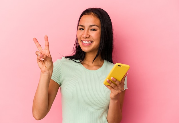 Joven venezolana sosteniendo un teléfono móvil aislado sobre fondo rosa mostrando el número dos con los dedos.