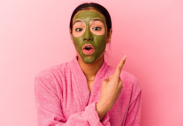 Joven venezolana vistiendo una bata de baño y una máscara facial aislada sobre fondo rosa apuntando hacia el lado