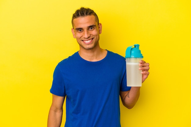 Joven venezolano bebiendo un batido de proteínas aislado en la pared amarilla feliz, sonriente y alegre.