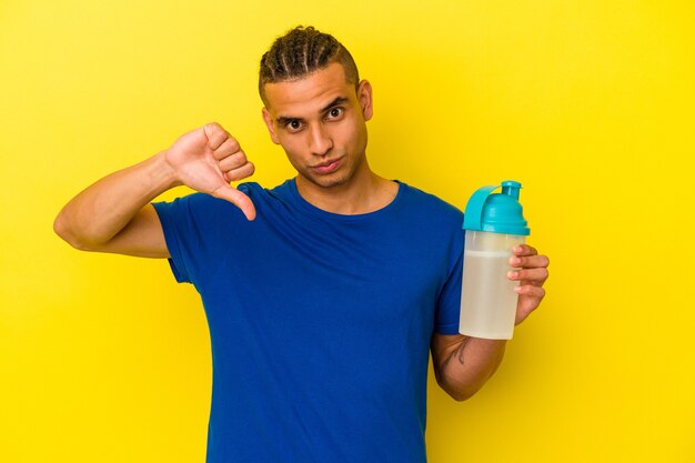 Joven venezolano bebiendo un batido de proteínas aislado sobre fondo amarillo mostrando un gesto de aversión, pulgares hacia abajo. Concepto de desacuerdo.