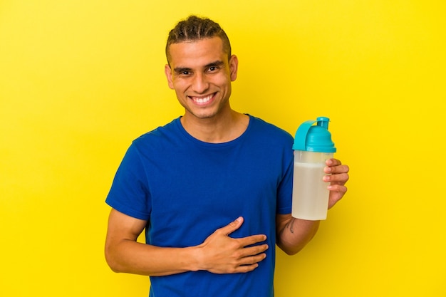 Joven venezolano bebiendo un batido de proteínas aislado sobre fondo amarillo riendo y divirtiéndose.