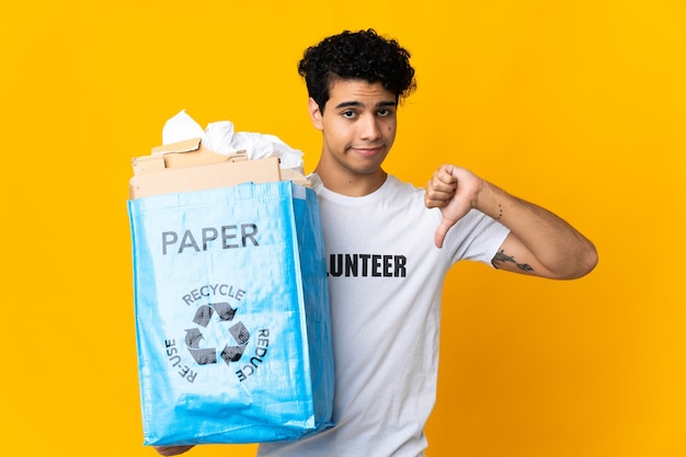 Joven venezolano sosteniendo una bolsa de reciclaje llena de papel para reciclar mostrando el pulgar hacia abajo con expresión negativa