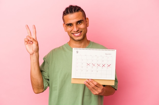 Joven venezolano sosteniendo un calendario aislado sobre fondo rosa alegre y despreocupado mostrando un símbolo de paz con los dedos.