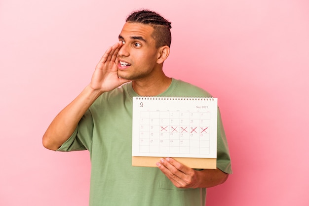 Joven venezolano sosteniendo un calendario aislado sobre fondo rosa gritando y sosteniendo la palma cerca de la boca abierta.