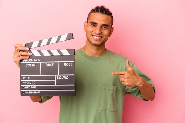 Joven venezolano sosteniendo una claqueta aislada sobre fondo rosa persona apuntando con la mano a un espacio de copia de camisa, orgulloso y seguro