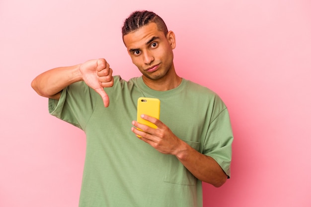 Joven venezolano sosteniendo un teléfono móvil aislado en la pared rosa mostrando un gesto de aversión, pulgares hacia abajo. Concepto de desacuerdo.