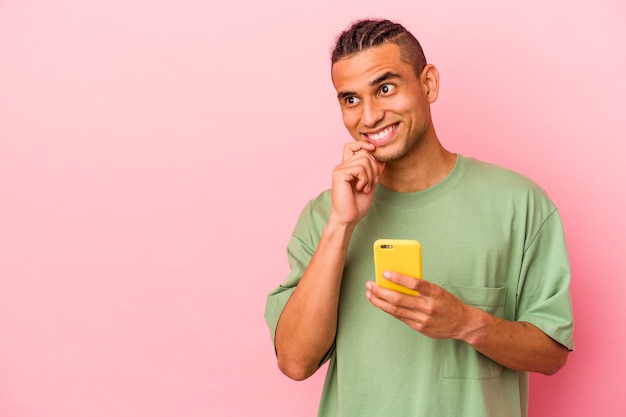 Joven venezolano sosteniendo un teléfono móvil aislado en la pared rosa relajado pensando en algo mirando un espacio de copia.