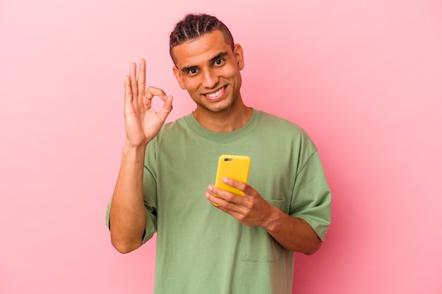 Joven venezolano sosteniendo un teléfono móvil aislado sobre fondo rosa alegre y confiado mostrando gesto ok.