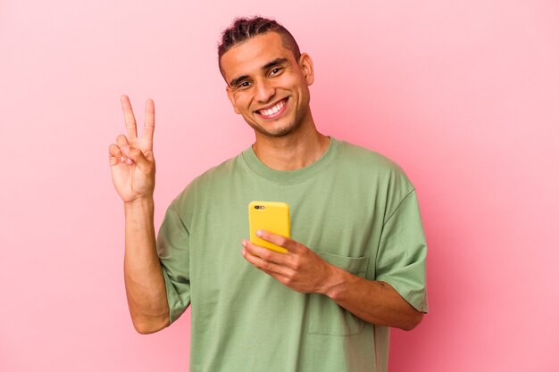 Joven venezolano sosteniendo un teléfono móvil aislado sobre fondo rosa alegre y despreocupado mostrando un símbolo de paz con los dedos.