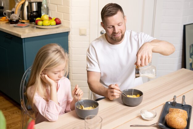 Joven vertiendo leche fresca en un tazón de porcelana gris con muesli o copos de maíz mientras desayuna con su linda hijita