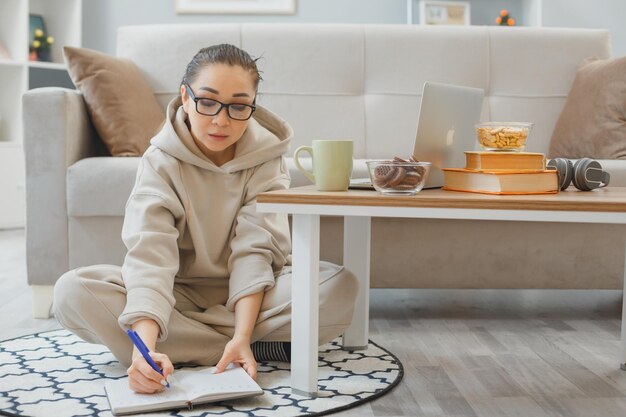 joven vestida de casa con anteojos sentada en un piso cerca de un sofá en el interior de su casa usando una laptop con entrenamiento en línea bebiendo café con confianza enfocada en una tarea