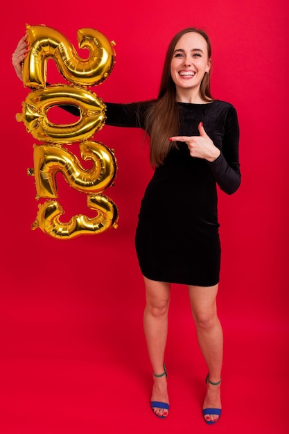 Una joven vestida festivamente con un vestido negro y tacones sostiene los números del nuevo año 2023 en un fondo rojo