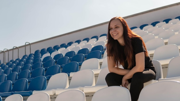 Una joven vestida de negro con el pelo largo está sentada sola en las gradas de un estadio y viendo un partido deportivo bajo el sol abrasador