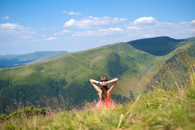 Joven vestida de rojo descansando en un campo de hierba verde en un cálido día soleado en las montañas de verano disfrutando de la vista de la naturaleza