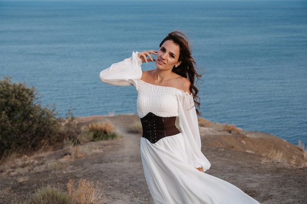 Una joven con un vestido blanco posa en el acantilado de la montaña Detrás de ella está el mar azul El vestido y el cabello ondean en el viento El concepto de libertad