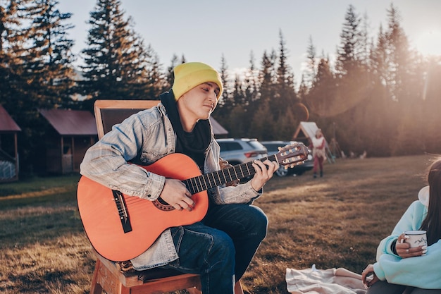 Un joven vestido con una chaqueta vaquera y sosteniendo una guitarra acústica tocando una melodía. El fondo del centro de recreación forestal y el sol brillante.