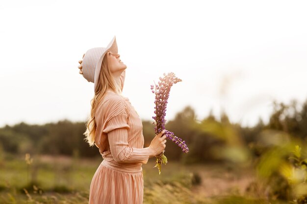 Foto una joven con un vestido largo y un sombrero camina por el campo con un ramo