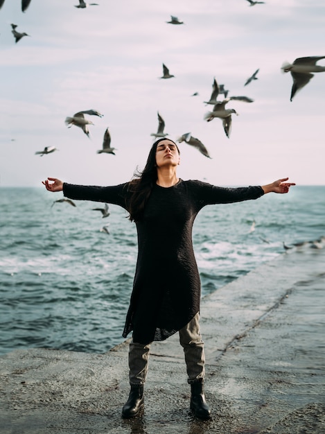 Una joven con un vestido negro y jeans está junto al mar con un fuerte viento.