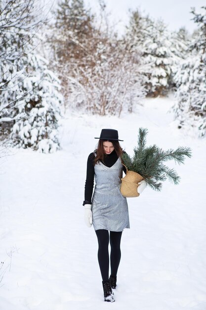 Una joven con un vestido plateado y un sombrero se para con la espalda en medio de un hermoso bosque de invierno