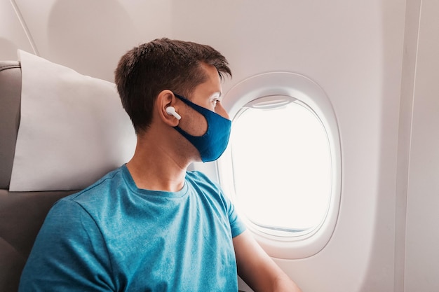 El joven viaja en avión usa una máscara facial durante la pandemia de Covid19 y escucha música con auriculares y mira por la ventana