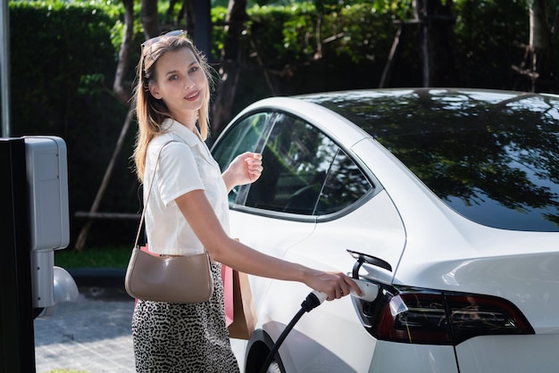Una joven viaja con un coche eléctrico EV cargando en un jardín verde y sostenible al aire libre en una ciudad en verano Estilo de vida de sostenibilidad urbana con energía verde, limpia y recargable del interior de un vehículo eléctrico BEV